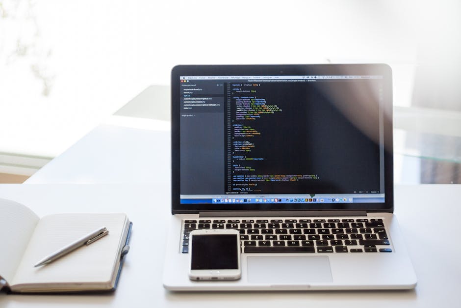 A silver laptop displaying a code editor with lines of colorful programming code on the screen, placed on a white desk inside a well-lit room. To the left of the laptop, there is an open notebook with