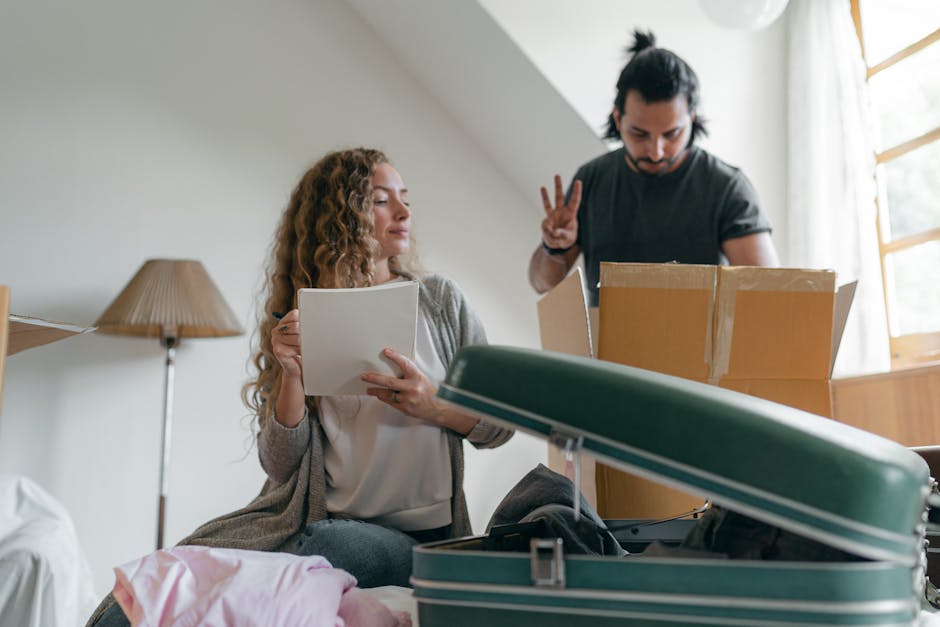A young woman with curly hair sits on a bed inside a room, holding a document or checklist, while a man with long dark hair stands nearby, showing a peace sign with his fingers. The room contains a gr