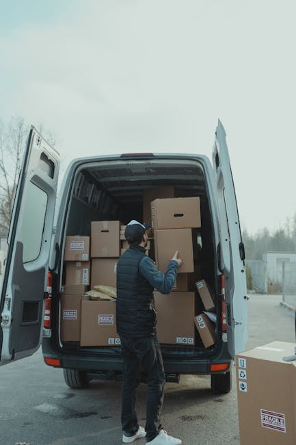 A man with a serious expression, wearing a white polo shirt and a black cap, stands inside the rear of a moving van with the doors open. The van's interior is filled with neatly stacked cardboard boxe